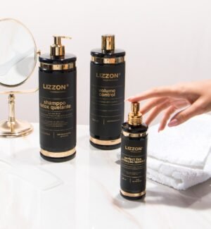 Três garrafas pretas e douradas da Kit Liso Blindado Promo ficam em um balcão branco ao lado de um espelho redondo e toalhas dobradas, enquanto uma mão alcança a menor garrafa na frente.