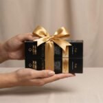 Duas mãos seguram duas caixas retangulares pretas empilhadas e amarradas com uma fita de cetim dourada. As caixas apresentam o nome do produto, "Colorizze em dobro: coloração creme permanente", em texto dourado. O plano de fundo é neutro e simples.