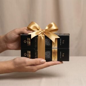 Duas mãos seguram duas caixas retangulares pretas empilhadas e amarradas com uma fita de cetim dourada. As caixas apresentam o nome do produto, "Colorizze em dobro: coloração creme permanente", em texto dourado. O plano de fundo é neutro e simples.