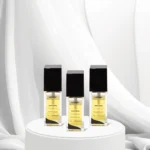 Três frascos do perfume Lizzon Ouro Inka com tampas pretas e rótulos dourados são exibidos em um pedestal redondo branco, em um fundo de tecido branco macio, mostrando a elegância do estilo característico de Lizzon.