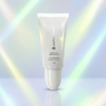 Um tubo branco com o rótulo Sérum Perfect Brows 15 ml está em pé em uma superfície refletora contra um fundo suave em tons de arco-íris.